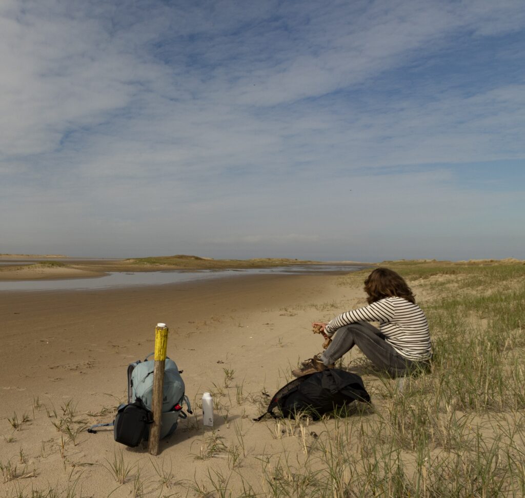 Adriënne Naron zittend op in vlakke duinen met een rugzak uitkijkend op het water